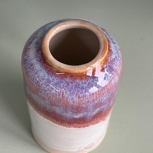 Puede incluir: Un jarrón de cerámica hecho a mano con forma cilíndrica. La sección superior presenta una mezcla de tonos morados, rojos y azules, mientras que la mitad inferior es de color blanco crema. El jarrón tiene un acabado liso y brillante y un borde de color neutro.