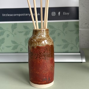Puede incluir: Un difusor de caña de cerámica hecho a mano con un acabado rústico y texturizado. El jarrón presenta una mezcla de tonos marrones y rojos, con una base crema. Varias cañas de madera se insertan en el jarrón. El fondo muestra "Little Acorn Pottery".