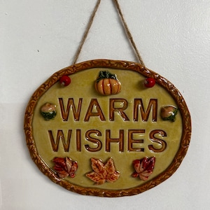 May include: A round, tan ceramic sign with a brown rope for hanging. The sign has the words "Warm Wishes" in brown lettering. There are small, decorative autumn-themed accents around the words, including pumpkins, acorns, and leaves.