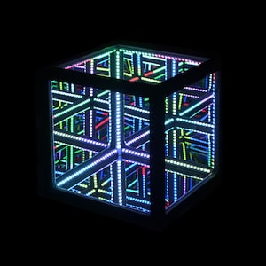 May include: A black cube with a mirrored interior, lit by a network of colourful LED lights. The lights create an illusion of infinite depth, with colours including blue, green, and red. The cube is a modern art piece.