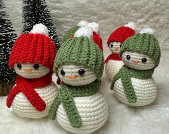 Puede incluir: Cuatro adornos de muñeco de nieve de crochet con cuerpos blancos, sombreros rojos y verdes y bufandas a juego. Los muñecos de nieve están sentados sobre una superficie blanca y esponjosa.