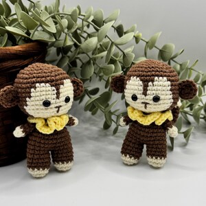 May include: Two handmade crochet monkey dolls. Each monkey has a brown body, head, and ears, with a cream-colored face and hands. They wear yellow ruffled collars. The monkeys are standing in front of green foliage.