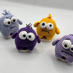May include: Four handmade crochet monster toys in blue, purple, and yellow. Each has large white eyes with black pupils, small arms, and a stitched mouth. The toys are sitting on small, round bases.