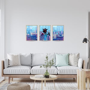 May include: Three framed prints of a comic book style illustration of Spider-Man characters swinging through a city skyline. The prints are hung on a white wall above a light-colored couch in a living room.
