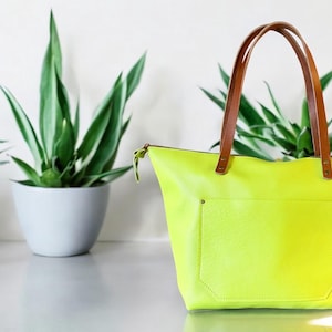 May include: A bright yellow leather tote bag with brown leather handles. The bag has a zipper closure and a large front pocket.