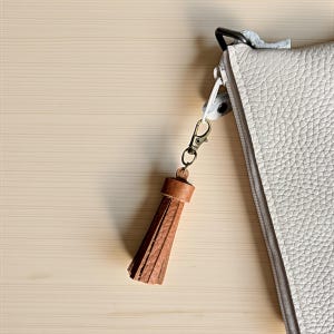 May include: A light gray leather clutch with a zipper closure and a brown leather tassel attached to a gold-colored clasp. The clutch has a textured surface and is set against a light wood background.
