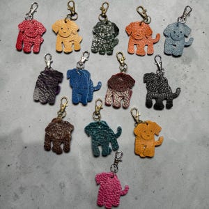 May include: A collection of leather dog-shaped keychains in various colors, including red, yellow, green, orange, blue, and gray. Each keychain has a metal clasp for attaching to keys or bags. The keychains are arranged on a gray surface.