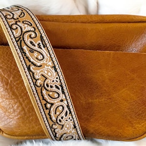 May include: A brown leather crossbody bag with a paisley patterned strap. The bag has a zipper closure and a flap.