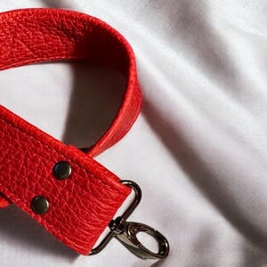 May include: A red leather strap with a silver hook. The strap has a textured surface and two silver rivets.