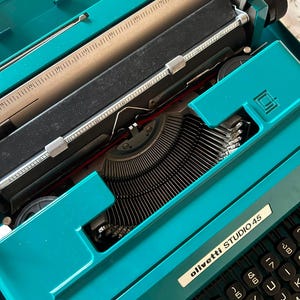 Puede incluir: Una máquina de escribir Olivetti Studio 45 de color turquesa. Tiene un teclado negro y un mecanismo de alimentación de papel. El texto "olivetti STUDIO 45" es visible en una etiqueta blanca. La máquina está abierta, mostrando la mecánica interna.