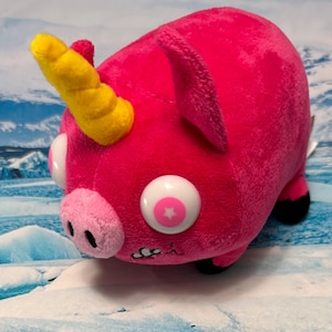 Peut inclure: Un cochon en peluche avec une corne jaune, un groin rose et de grands yeux blancs avec des centres en forme d'étoile rose. Le cochon a un corps rose vif, des sabots noirs et un design fantaisiste. Le jouet est conçu pour ressembler à un hybride cochon-licorne.