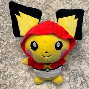 May include: Yellow plush toy with black ears and a red hooded sweatshirt. The toy has a white shirt with a Pokeball logo and a red X on the front.