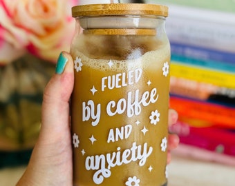 May include: A glass jar with a bamboo lid and a straw, filled with a brown liquid. The jar has a white graphic that says "Fueled by coffee and anxiety" with small white flowers and stars around the text.