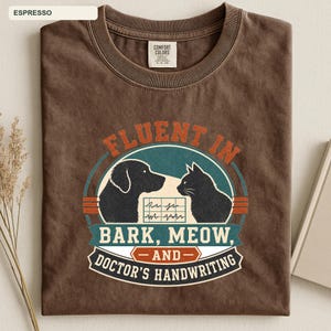 May include: A dark brown crew neck t-shirt featuring a graphic with a dog and a cat silhouette. The text reads FLUENT IN BARK, MEOW, AND DOCTOR'S HANDWRITING, with a central box showing a jagged line pattern. The shirt has a Comfort Colors label.