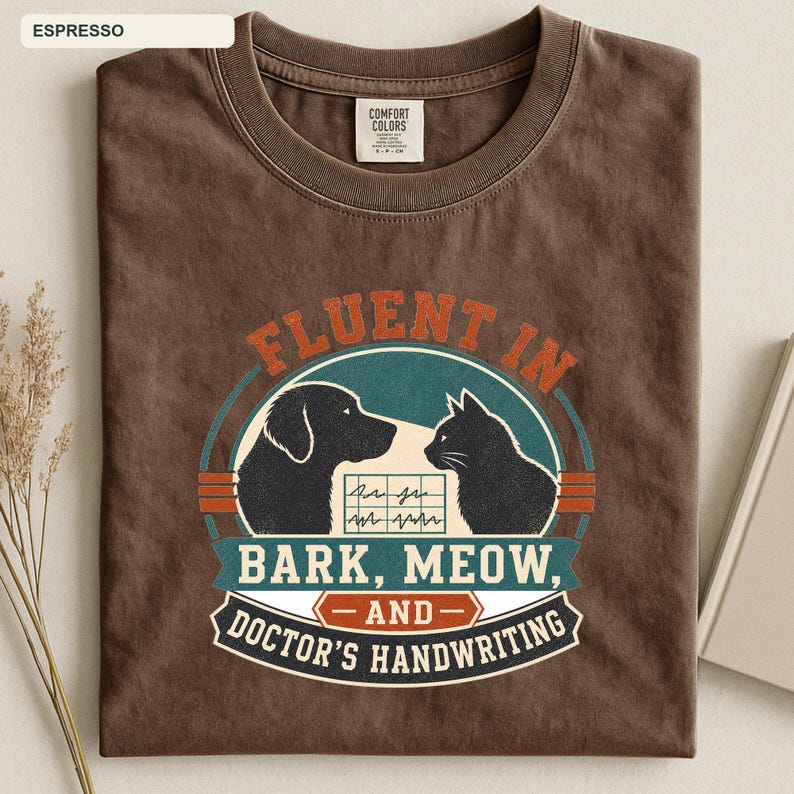May include: A dark brown crew neck t-shirt featuring a graphic with a dog and a cat silhouette. The text reads FLUENT IN BARK, MEOW, AND DOCTOR'S HANDWRITING, with a central box showing a jagged line pattern. The shirt has a Comfort Colors label.