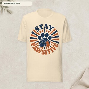 May include: A heather natural short-sleeve t-shirt featuring a circular graphic with dark blue and orange sunburst rays. The center displays a dark blue paw print containing silhouettes of a dog and a cat, with the text STAY PAWSITIVE.
