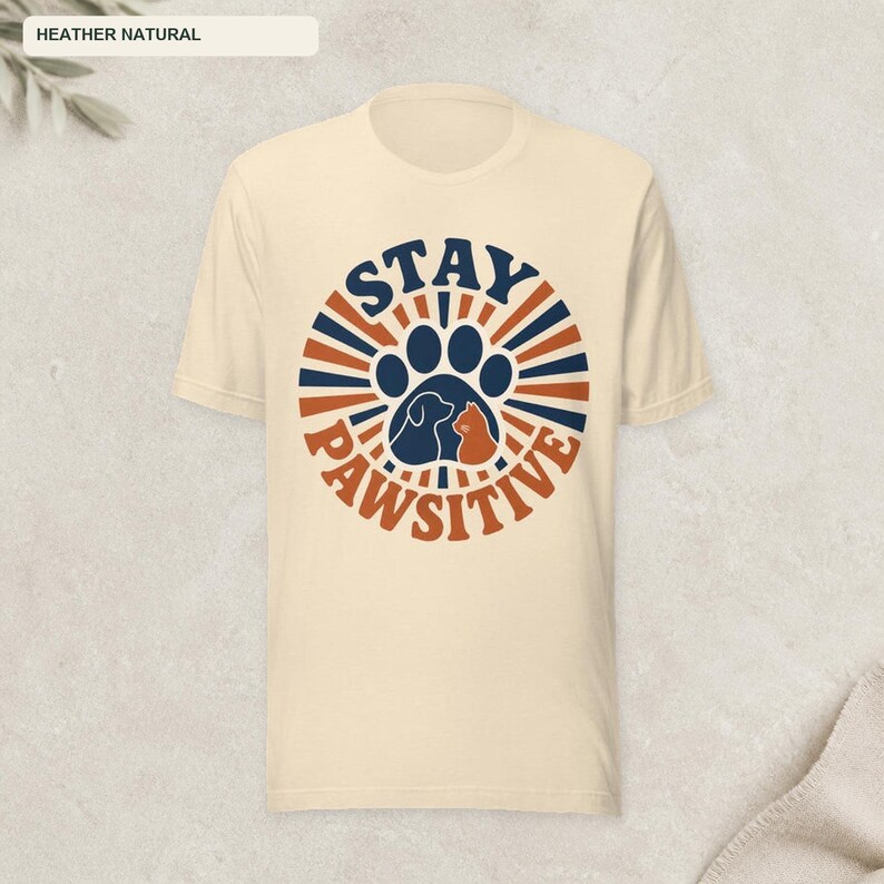 May include: A heather natural short-sleeve t-shirt featuring a circular graphic with dark blue and orange sunburst rays. The center displays a dark blue paw print containing silhouettes of a dog and a cat, with the text STAY PAWSITIVE.