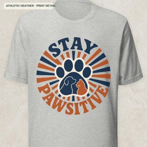 May include: An athletic heather gray short-sleeve t-shirt featuring a circular graphic with the text STAY PAWSITIVE. The design includes a large paw print containing silhouettes of a dog and a cat, surrounded by alternating navy blue and orange rays.