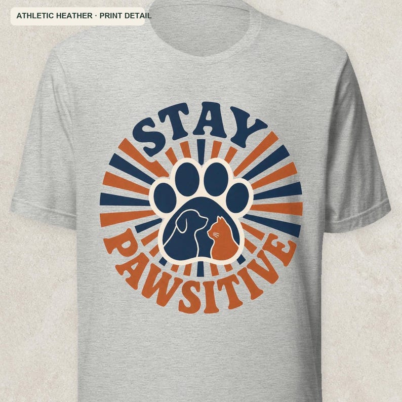 May include: An athletic heather gray short-sleeve t-shirt featuring a circular graphic with the text STAY PAWSITIVE. The design includes a large paw print containing silhouettes of a dog and a cat, surrounded by alternating navy blue and orange rays.