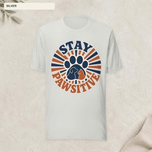 May include: A silver-colored short-sleeve t-shirt featuring a circular graphic with dark blue and orange sunburst rays. The center displays a large paw print containing silhouettes of a dog and a cat, with the text STAY PAWSITIVE arched around.