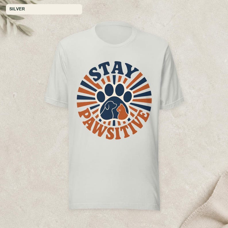May include: A silver-colored short-sleeve t-shirt featuring a circular graphic with dark blue and orange sunburst rays. The center displays a large paw print containing silhouettes of a dog and a cat, with the text STAY PAWSITIVE arched around.