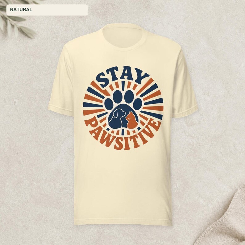 May include: A natural-colored t-shirt featuring a circular graphic with dark blue and orange sunburst rays. In the center is a large dark blue paw print containing silhouettes of a dog and a cat, with the text STAY PAWSITIVE arched around it.