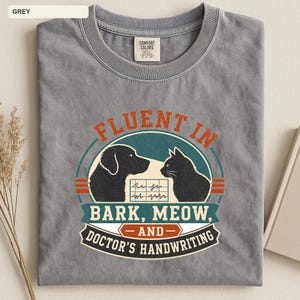 May include: A grey crew neck t-shirt featuring a graphic print of a dog and a cat silhouette facing each other. The text reads FLUENT IN BARK, MEOW, AND DOCTOR'S HANDWRITING, with a medical chart icon between the animals in a circular design.