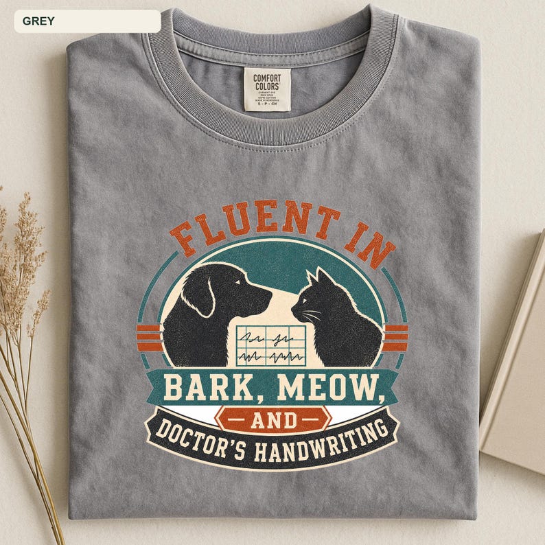 May include: A grey crew neck t-shirt featuring a graphic print of a dog and a cat silhouette facing each other. The text reads FLUENT IN BARK, MEOW, AND DOCTOR'S HANDWRITING, with a medical chart icon between the animals in a circular design.