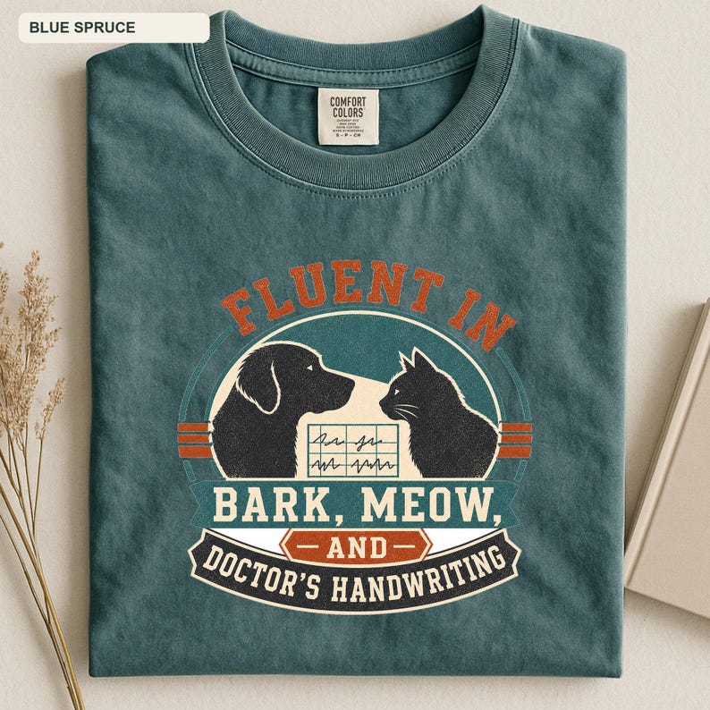 May include: A blue spruce t-shirt featuring a graphic of a dog and a cat facing each other with a medical chart in between. The text reads: FLUENT IN BARK, MEOW, AND DOCTOR'S HANDWRITING. The shirt has a crew neck and a Comfort Colors brand label.