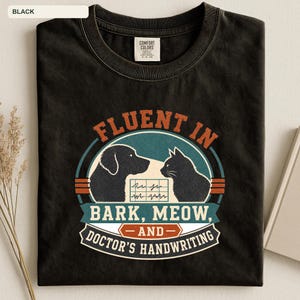 May include: A black crew neck t-shirt featuring a graphic print with a dog and a cat silhouette facing each other. The text reads FLUENT IN BARK, MEOW, AND DOCTOR'S HANDWRITING, with a medical chart symbol between the animals in a vintage color palette.