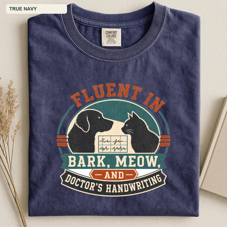 May include: A true navy short-sleeve t-shirt featuring a graphic print of a dog and a cat silhouette. The text reads: FLUENT IN BARK, MEOW, AND DOCTOR'S HANDWRITING. A small grid with wavy lines is positioned between the animal silhouettes.