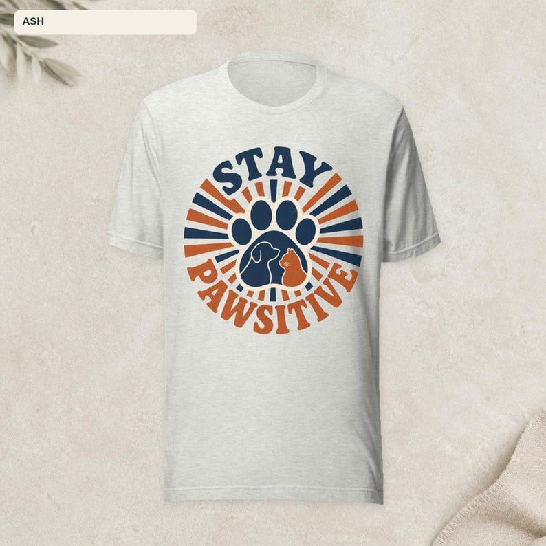 May include: An ash gray short-sleeve t-shirt featuring a circular graphic with radiating blue and orange lines. The design includes a central paw print containing silhouettes of a dog and a cat, with the text STAY PAWSITIVE printed around it.