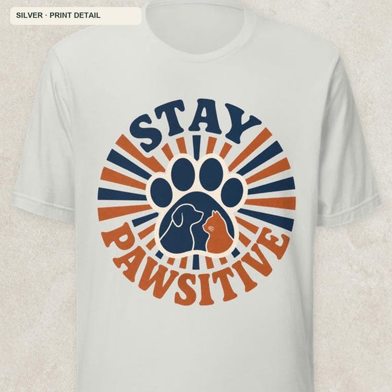 May include: A light gray short-sleeve t-shirt featuring a circular graphic with alternating dark blue and orange sunburst rays. The center contains a large dark blue paw print silhouette with a dog and cat profile, and the text STAY PAWSITIVE.