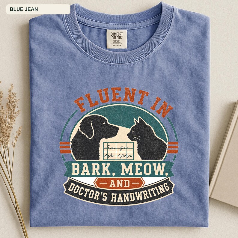 May include: A blue jean colored t-shirt featuring a graphic with a black dog and a black cat silhouette facing each other. Between them is a medical chart icon. The text reads Fluent In Bark, Meow, And Doctor's Handwriting in orange and white.