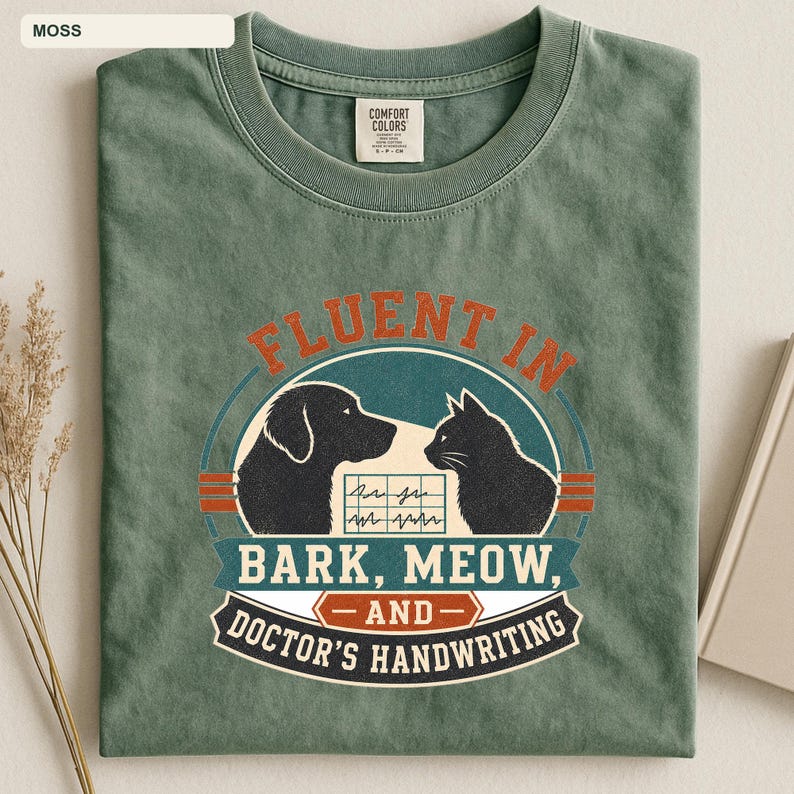 May include: A moss green crew neck t-shirt featuring a graphic of a dog and a cat facing each other. The text reads "FLUENT IN BARK, MEOW, AND DOCTOR'S HANDWRITING" with a small EKG line graphic between the animals. The shirt has a Comfort Colors label.