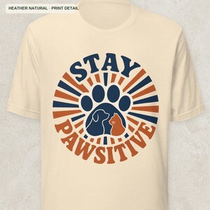 May include: A heather natural short-sleeve t-shirt featuring a circular graphic with dark blue and orange sunburst rays. The center contains a dark blue paw print silhouette with a dog and cat profile, and the text STAY PAWSITIVE.