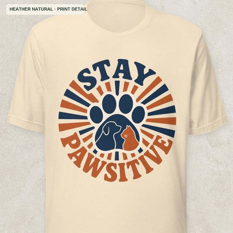May include: A heather natural short-sleeve t-shirt featuring a circular graphic with dark blue and orange sunburst rays. The center contains a dark blue paw print silhouette with a dog and cat profile, and the text STAY PAWSITIVE.