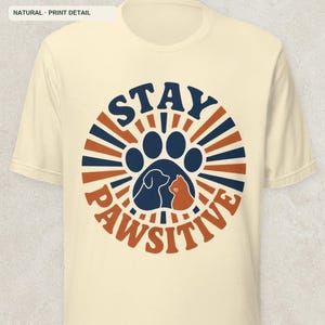 May include: A natural-colored short-sleeve t-shirt featuring a circular graphic with dark blue and orange sunburst rays. The center displays a large paw print containing silhouettes of a dog and a cat, with the text STAY PAWSITIVE in bold.