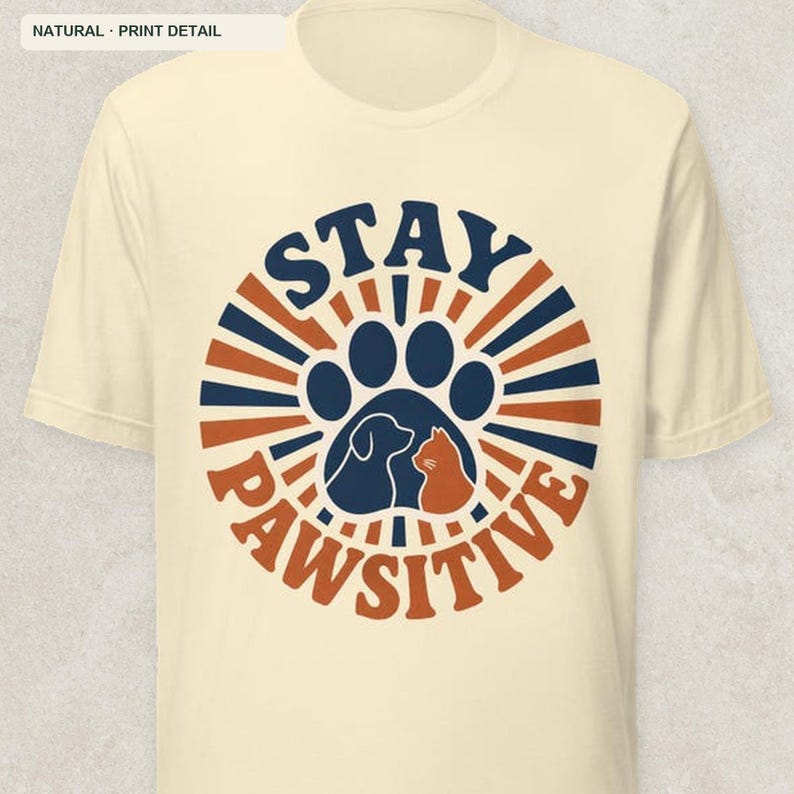 May include: A natural-colored short-sleeve t-shirt featuring a circular graphic with dark blue and orange sunburst rays. The center displays a large paw print containing silhouettes of a dog and a cat, with the text STAY PAWSITIVE in bold.