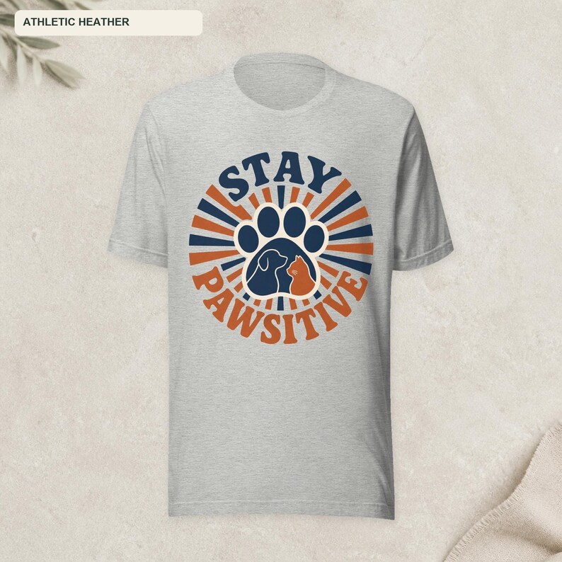 May include: An athletic heather gray short-sleeve t-shirt featuring a circular graphic with dark blue and orange sunburst rays. The center displays a large paw print containing silhouettes of a dog and a cat, with the text STAY PAWSITIVE.