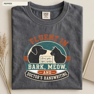 May include: A charcoal gray crew neck t-shirt featuring a graphic print of a dog and a cat facing each other. The text reads FLUENT IN BARK, MEOW, AND DOCTOR'S HANDWRITING, with a central EKG line graphic. The shirt has a Comfort Colors brand label.