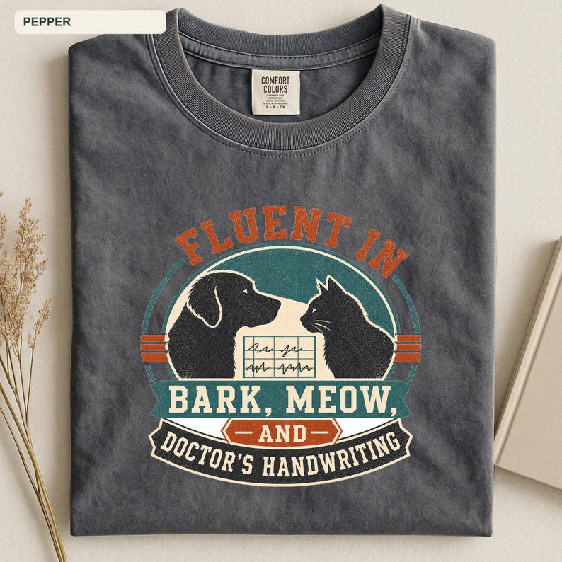 May include: A charcoal gray crew neck t-shirt featuring a graphic print of a dog and a cat facing each other. The text reads FLUENT IN BARK, MEOW, AND DOCTOR'S HANDWRITING, with a central EKG line graphic. The shirt has a Comfort Colors brand label.