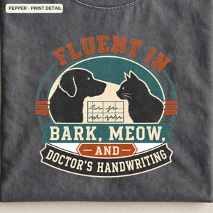 May include: A charcoal gray t-shirt featuring a graphic with a black dog and cat silhouette facing each other. Between them is a medical chart icon. The text reads: FLUENT IN BARK, MEOW, AND DOCTOR'S HANDWRITING. The design uses teal, orange, and white colors.
