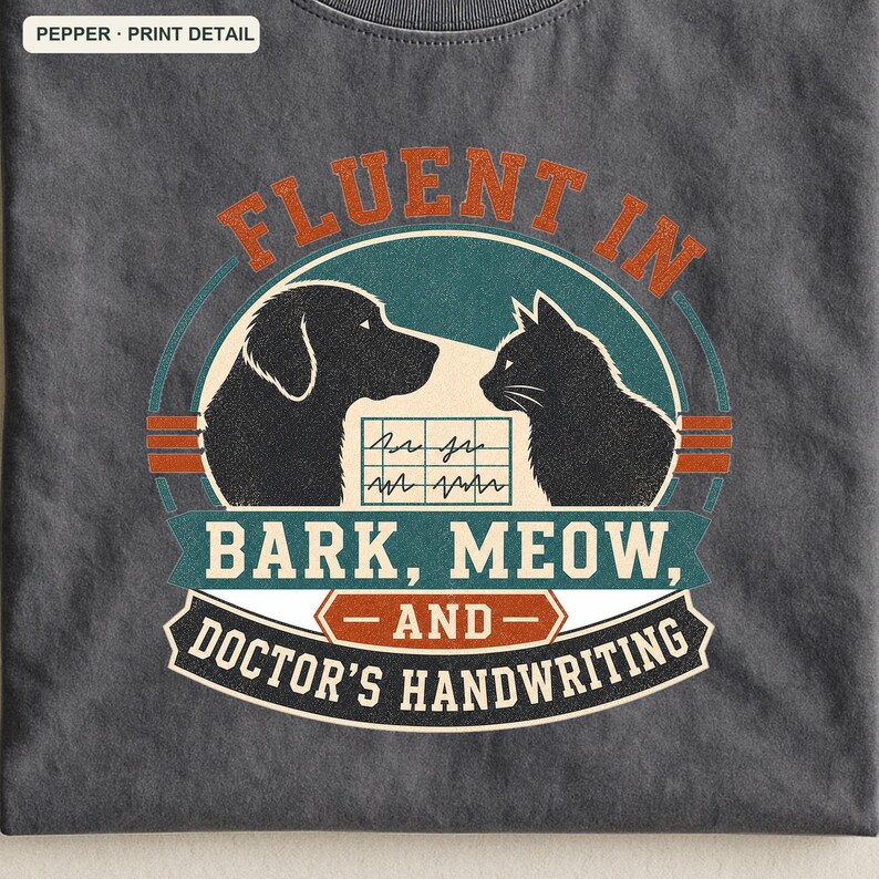 May include: A charcoal gray t-shirt featuring a graphic with a black dog and cat silhouette facing each other. Between them is a medical chart icon. The text reads: FLUENT IN BARK, MEOW, AND DOCTOR'S HANDWRITING. The design uses teal, orange, and white colors.