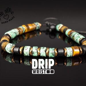 Peut inclure: Un bracelet de perles composé de perles vertes, noires et marron avec des accents dorés. Le bracelet est monté sur un cordon noir. Le texte "DRIP WRIST" est affiché en lettres blanches. Fait à la main.