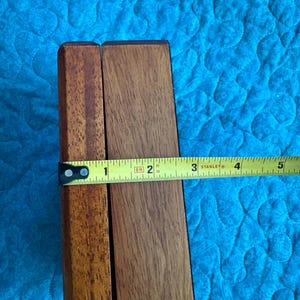K&ouml;nnte beinhalten: Zwei Holzst&uuml;cke, vermutlich von einem M&ouml;belst&uuml;ck, werden mit einem Ma&szlig;band gezeigt. Das Band zeigt eine Breite von etwa 5 cm an. Das Holz hat einen warmen, braunen Ton, und der Hintergrund ist ein blaues Steppstoff.