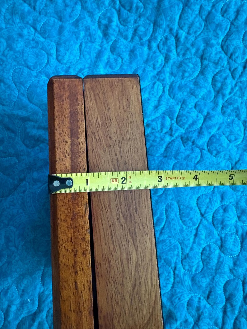 K&ouml;nnte beinhalten: Zwei Holzst&uuml;cke, vermutlich von einem M&ouml;belst&uuml;ck, werden mit einem Ma&szlig;band gezeigt. Das Band zeigt eine Breite von etwa 5 cm an. Das Holz hat einen warmen, braunen Ton, und der Hintergrund ist ein blaues Steppstoff.