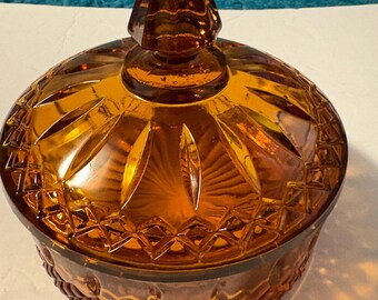 May include: Amber-colored glass candy dish with a diamond-cut pattern and a pointed lid. The dish has a round base and a decorative design, perfect for storing small items or displaying as a decorative piece. The glass has a warm, inviting hue.
