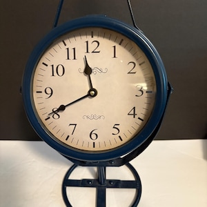 May include: A blue metal clock with a white face and black numerals. The clock has a handle and a decorative base. The hands point to approximately 11:58. The clock is a vintage style.