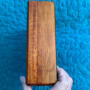K&ouml;nnte beinhalten: Eine rechteckige Holzkiste mit Scharnierdeckel, aus rotbraunem Holz gefertigt. Die Kiste wird in einer Hand vor einem blauen, gesteppten Hintergrund gehalten. Die Holzmaserung ist sichtbar und verleiht dem Design Textur.